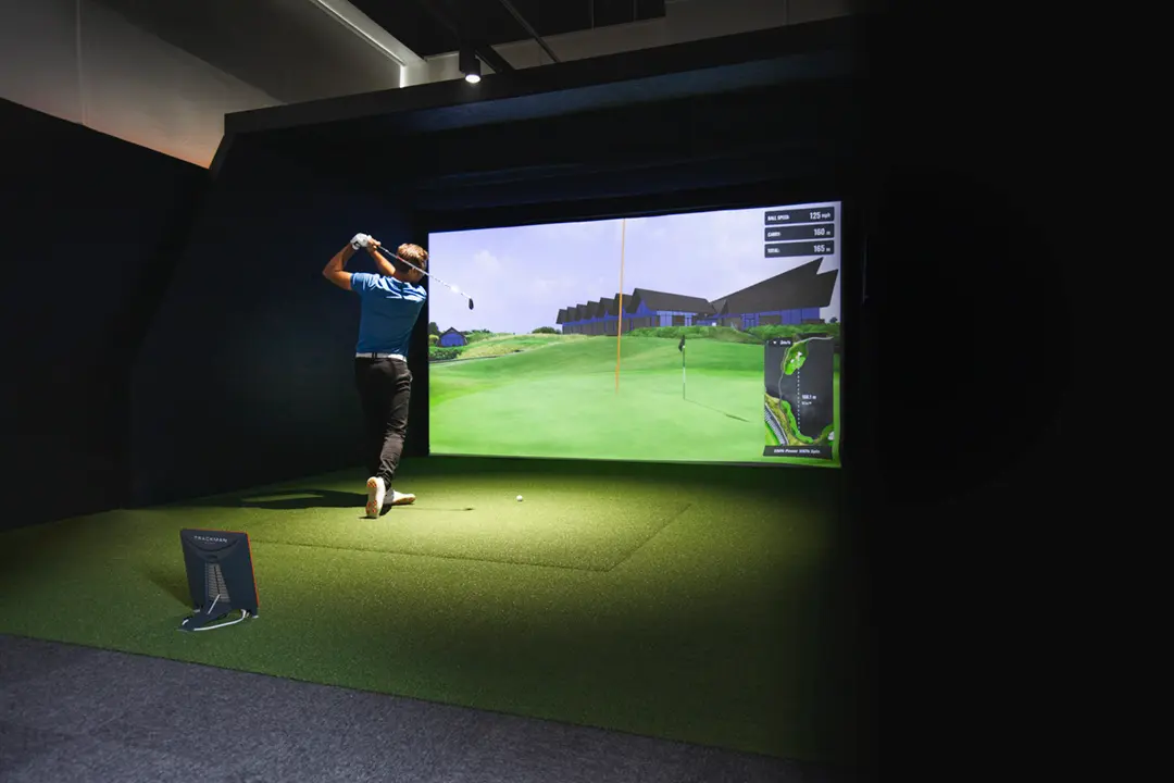 Golfer using a commercial indoor golf simulator in a clubhouse environment with large projection screen and launch monitor.