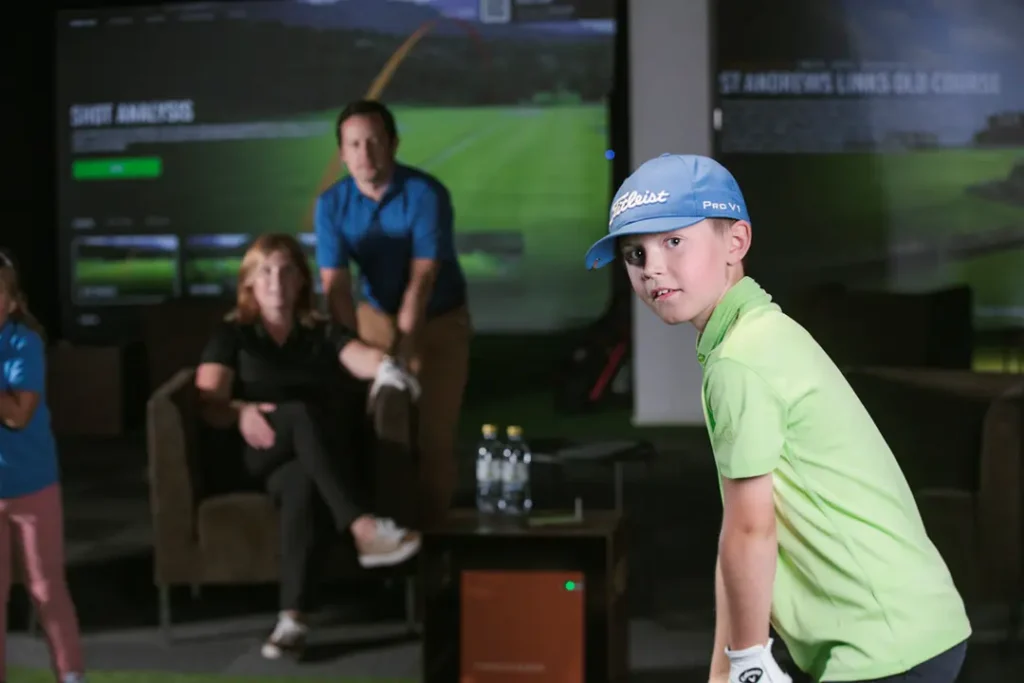 Family using a golf simulator in a clubhouse setting with child playing indoor golf and spectators watching.