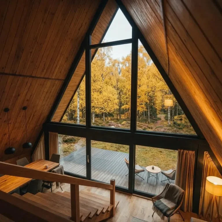 Interior of The Arla A-frame lodge by Heritage Lodges showing timber-lined walls, staircase, and large triangular window overlooking woodland.